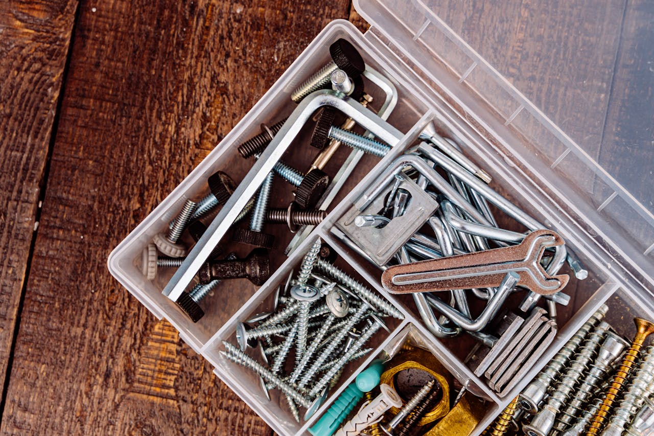 Top view of various screws and tools in a plastic container on wood surface.